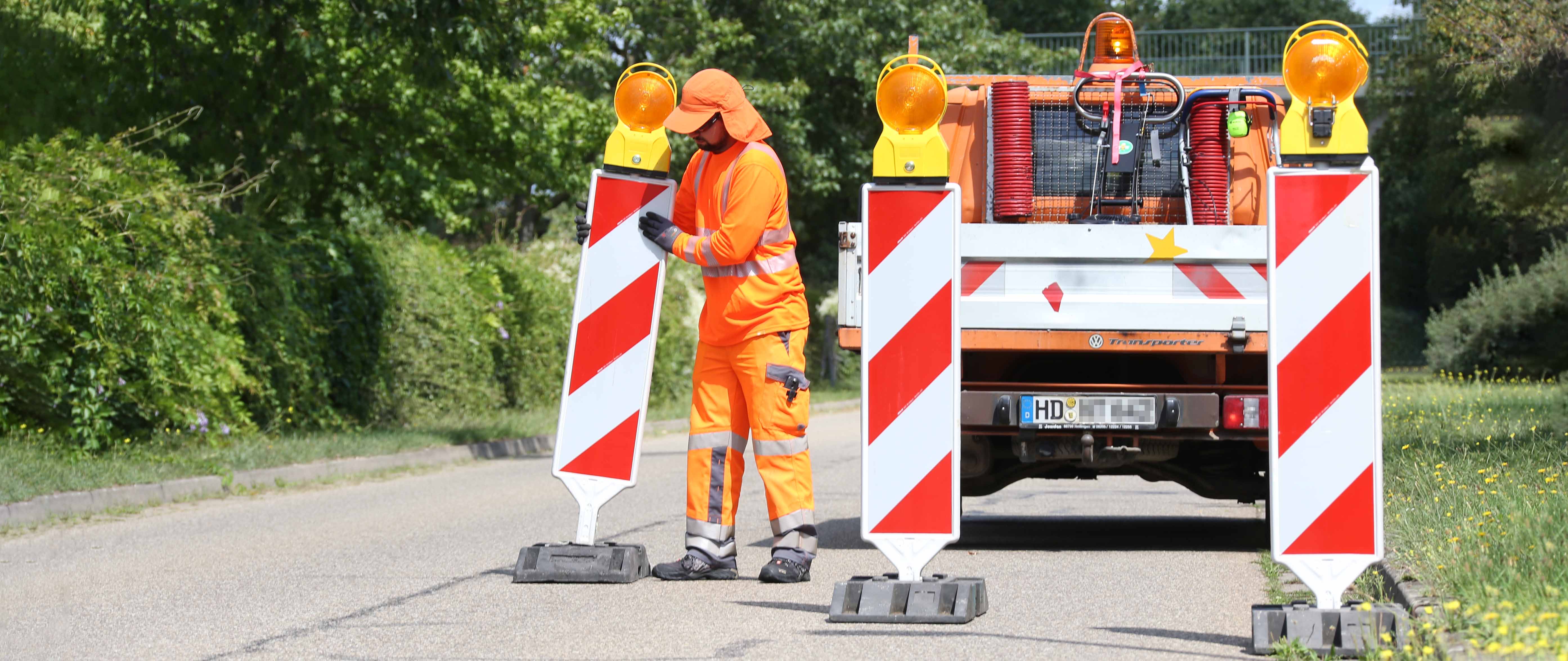 Ein Bauarbeiter in orangefarbener Schutzkleidung steht vor einem Fahrzeug mit Warnmarkierungen auf einer Straße. Er ist von Absperrbaken umgeben, während er Arbeiten im Straßenbereich durchführt.
