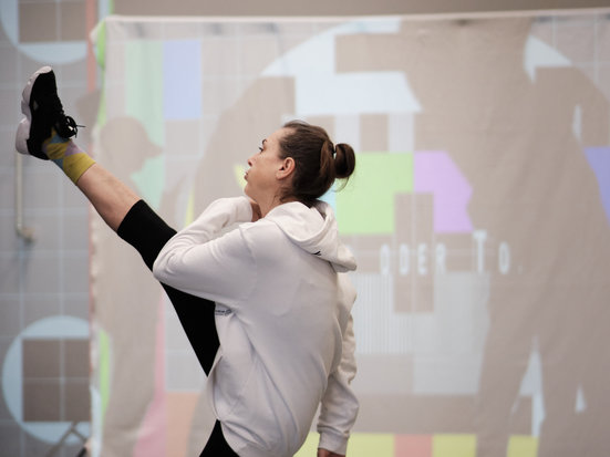 Eine Person in heller Kleidung führt in einem Raum eine dynamische Tanzbewegung aus und hebt dabei ein Bein hoch. Im Hintergrund sind unscharfe Projektionen oder bunte Formen an der Wand zu sehen.