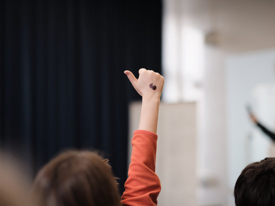 Eine Person im Publikum hebt die Hand in einer Faust, mit dem Daumen nach links herausschauend, um sich zu melden. Im Vordergrund sind weitere Zuhörende unscharf zu sehen, während der Fokus auf der erhobenen Hand liegt.
