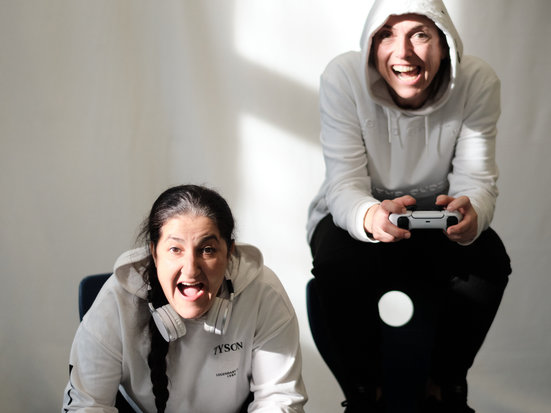 Zwei Personen sitzen entspannt in einem hellen Raum und lächeln in die Kamera. Eine sitzt vorne leicht nach vorn gebeugt, die andere dahinter auf einem Hocker, beide wirken freundlich und gelöst. Sie halten jeweils einen Controller in der Hand.