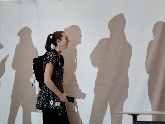 Eine Person geht an einer Wand entlang, auf der mehrere Schatten von Menschen zu sehen sind. Die Schatten wirken größer und verzerrt und erzeugen eine besondere Lichtstimmung.