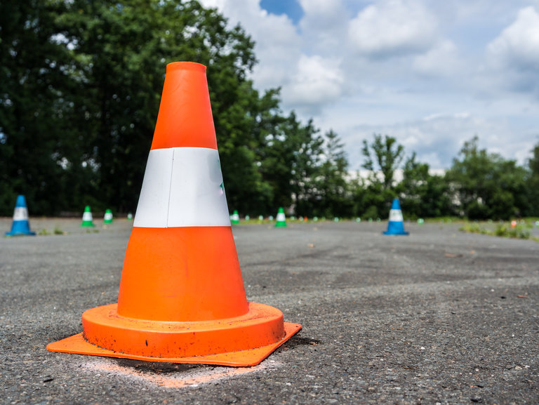 Ein orange-weißer Verkehrskegel steht im Vordergrund auf einer asphaltierten Fläche. Im Hintergrund sind weitere kleinere Kegel sowie Bäume und ein teilweise bewölkter Himmel zu sehen.