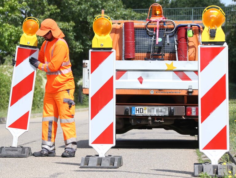 Ein Bauarbeiter in orangefarbener Schutzkleidung steht vor einem Fahrzeug mit Warnmarkierungen auf einer Straße. Er ist von Absperrbaken umgeben, während er Arbeiten im Straßenbereich durchführt.