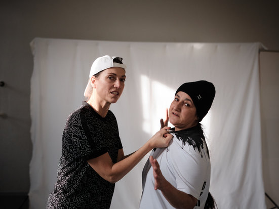 Zwei Personen stehen vor einem hellen Hintergrund. Die eine Person fasst der anderen Person an den Kragen des T-Shirts und schaut grimmig. Die andere Person hebt abwehrend ihre Hände.