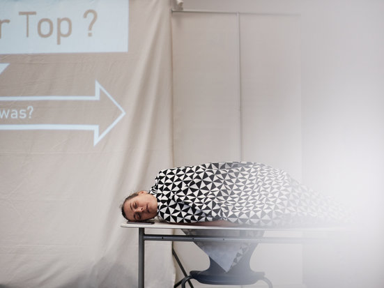 Eine Person mit gemustertem Oberteil liegt seitlich auf einem Tisch und schaut in die Kamera.