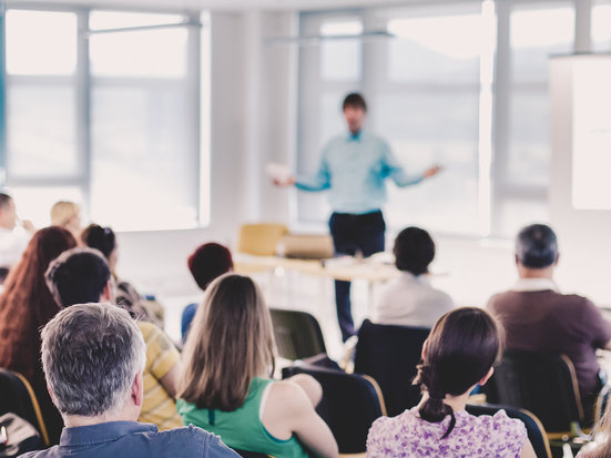 Eine Person steht vorne in einem hellen Raum und präsentiert vor einem sitzenden Publikum. Die Zuhörenden sind von hinten zu sehen und richten ihre Aufmerksamkeit auf die sprechende Person.